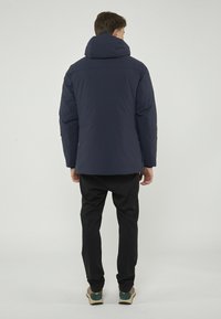 Giubbotto imbottito blu navy con colletto alto e cappuccio. Presenta una superficie liscia e texturizzata, tasche con zip e una vestibilità rilassata. Indossato con pantaloni neri.