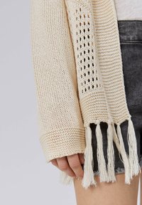 Hellbeige gestrickte Strickjacke mit offenem Muster, die Fransen am Saum und strukturierte Ärmel aufweist, vor grauem Hintergrund.