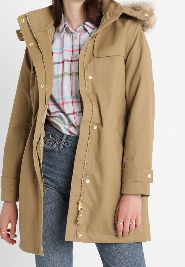 Kvinna iklädd en tanfärgad huva jacka över en flerfärgad rutad skjorta och blå jeans, med ringar på sina fingrar.