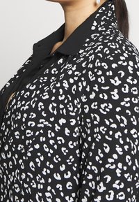 Chemise noire avec imprimé animalier blanc, dotée d'un col standard et d'une fermeture boutonnée sur le devant. Tissu léger avec une apparence texturée.