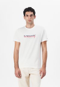 Hvit bomull T-skjorte med rund hals, med en blå og rød "tommy" logo over "JEANS" i matchende farger. Korte ermer, simpelt design.