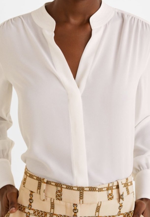 Femme portant un chemisier blanc à manches longues avec un col en V et un pantalon beige avec un motif imprimé chaîne dorée.