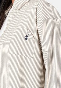 Stribet skjorte i beige og marineblå. Har en venstre brystlomme med en broderet detalje og en krave med løst, tekstureret stof.