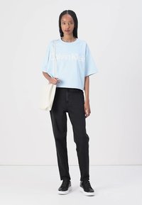 Ljust blå cropped t-shirt med vit text "Calvin Klein", tillsammans med svarta jeans och svarta sneakers, håller en krämfärgad tote-väska.