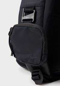Melnas neilona mugursomas detaļas, kurā ir ar rāvējslēdzēju aizverama kabata ar strukturētu virsmu un plastmasas sprādze. Nike logo eleganti attēlots.