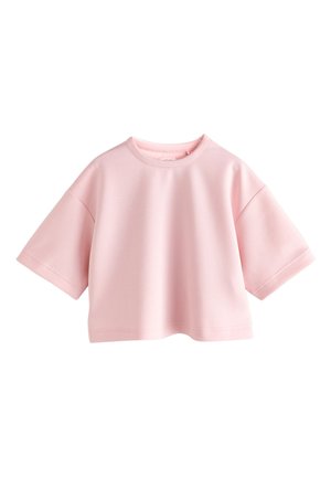 Jong meisje met lang haar, een losse roze top, wijde blauwe spijkerbroek en witte sandalen, staand tegen een effen achtergrond.