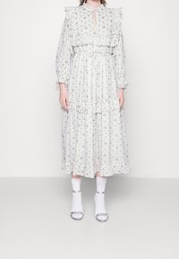 Valkoinen kukkamekko, jossa on röyhelökaulus ja -hihat, kerroksellinen muotoilu, vyötäröltä kiristetty, yhdistetty valkoisten sukkien ja hopeisten sandaalien kanssa.