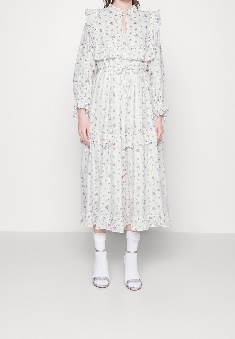 Bílé květinové šaty s volánkovým výstřihem a rukávy, vrstvený design, stahovaný pas, v kombinaci s bílými ponožkami a stříbrnými sandály.