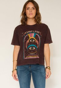 Femme portant un T-shirt marron avec un design d'œil cosmique coloré et de lune accompagné du texte "Astro Love", associé à un jean bleu et à des bracelets.