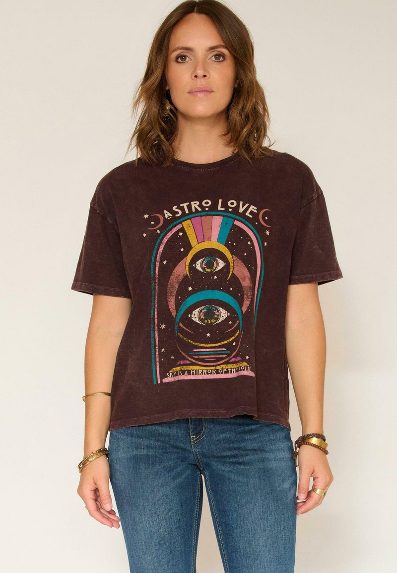 Femme portant un T-shirt marron avec un design d'œil cosmique coloré et de lune accompagné du texte "Astro Love", associé à un jean bleu et à des bracelets.