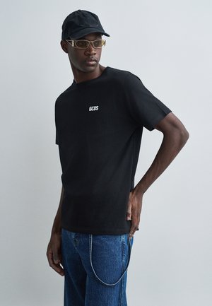 T-shirt nera con maniche corte e logo "GCDS" bianco sul petto, abbinata a jeans in denim blu e un cappellino nero, con occhiali da sole con montatura dorata.