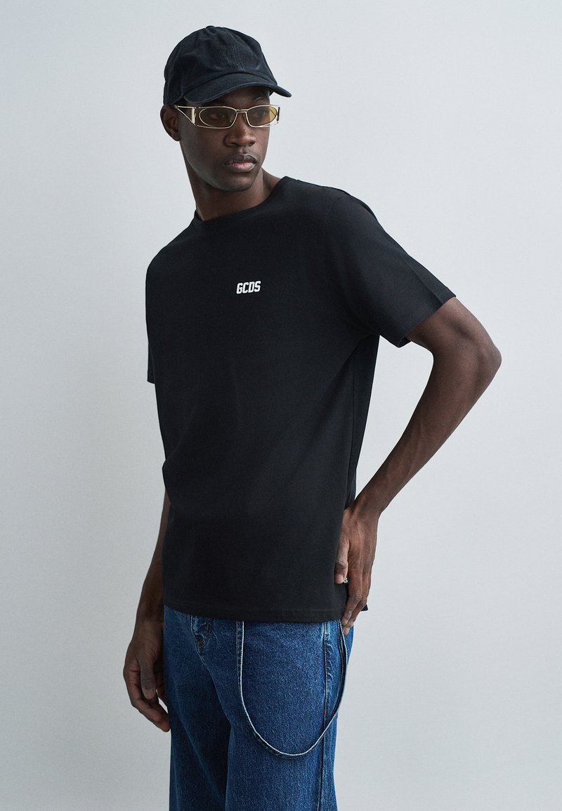 Čierne tričko s krátkymi rukávmi a bielym logom "GCDS" na hrudi, v kombinácii s modrými džínsami a čiernym klobúkom, ktorý má zlaté slnečné okuliare.