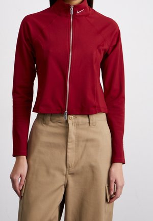 Femme portant une veste rouge ajustée zippée avec un col montant et un pantalon beige taille haute avec des poches sur les côtés.