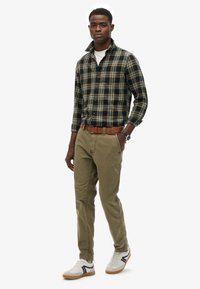 Geblokte button-up overhemd in donkergroen, beige en geel, gecombineerd met olijfgroene chinos en witte sneakers met zwarte accenten. Brown riem.