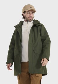 Grüne wasserdichte Jacke mit großen Taschen, Reißverschluss und verstellbarer Kapuze, über einer weißen Fleecejacke getragen. Darunter helle Cargo-Hosen.