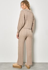 Beżowy sweter w splot kablowy i pasujące spodnie, z prążkowanymi mankietami, luźnym krojem i miękką, textured wykończeniem. Buty to jasnobeżowe trampki na platformie.