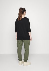 Top nero a tre quarti abbinato a pantaloni cargo verde oliva e sneakers beige, caratterizzato da una vestibilità rilassata e un design semplice.