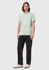 T-shirt verde menta chiaro a maniche corte, scollatura rotonda, abbinata a jeans di denim neri e sneakers bianche, in piedi contro uno sfondo semplice.