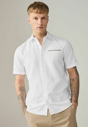 Young man with short blond hair wearing a white short-sleeve button-up shirt and beige pants, showing tattoos on both forearms.