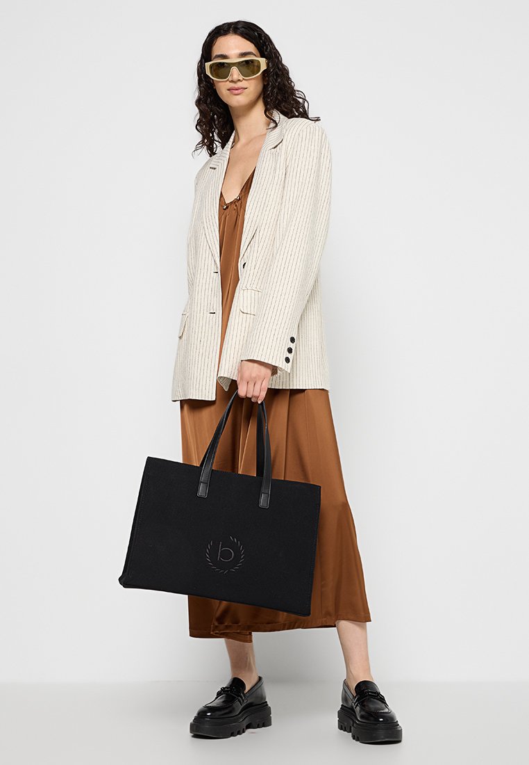Borsa a tracolla nera con logo in rilievo, texture in tessuto, manici lunghi; il modello indossa un blazer beige a righe sottili sopra un vestito marrone, abbinato a scarpe nere.