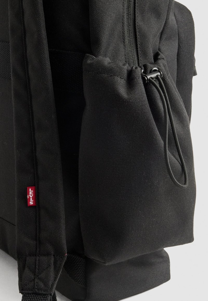 Schwarzer Stoff-Rucksack mit seitlichem Fach, verstellbarem Zugverschluss und rotem Levi's-Logo-Tag am Schultergurt vor weißem Hintergrund.