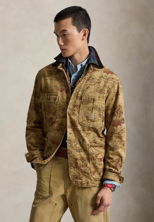 Uomo che indossa una giacca beige con motivo sopra una camicia di jeans blu, pantaloni beige, collana colorata di perline e braccialetto rosso, guarda di lato.
