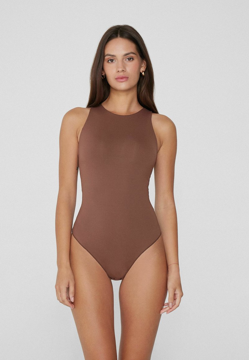 Mujer con cabello largo y oscuro, vistiendo un bodysuit sin mangas de color marrón con cuello alto, de pie contra un fondo gris claro liso.