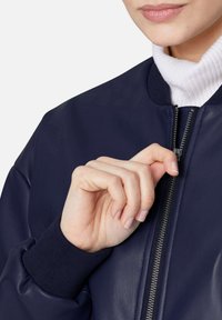 Marine bomberjack met leren stof en geribbelde manchetten, voorzien van een metalen ritssluiting en een witte coltrui eronder, waarmee het strakke ontwerp wordt benadrukt.