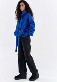 Hanorac supradimensionat albastru cu șnururi lungi și buzunar frontal, asortat cu blugi negri cu talie înaltă și adidași negri.