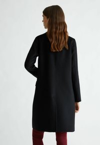 Cappotto nero con maniche lunghe e taglio dritto, con una cucitura posteriore e tasche laterali, abbinato a pantaloni bordeaux.