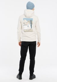 Felpa crema con una grafica di montagne da sci sulla schiena, indossata con pantaloni neri e un berretto di maglia azzurro chiaro, completata da scarpe nere.