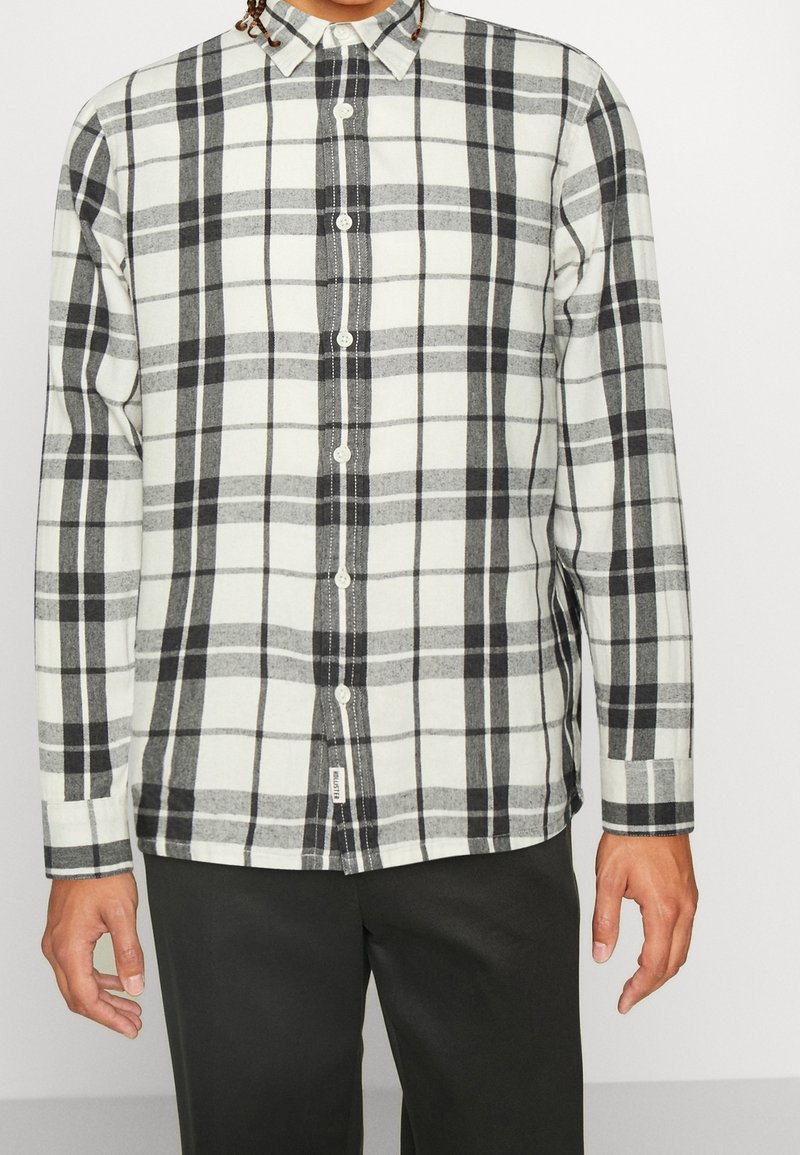 Mans långärmad skjorta i vitt med ett mönster i svart och grå rutigt. Tillverkad av mjukt tyg med en knappad krage och synlig sömnad.