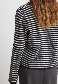 Veste en tricot à rayures noires et blanches avec des manches longues, tissu texturé et encolure ronde, associée à un pantalon gris.