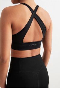 Svart sports-bh med kryssryggdesign, glatt tekstur og elastisk bånd. Logo sydd midt under båndet.