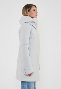 Donna con cappotto invernale grigio chiaro con cappuccio e jeans blu, in profilo su uno sfondo bianco uniforme.