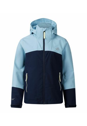 Jonge persoon draagt een blauw en marineblauw jack met marineblauwe korte broek, houdt een blauwgroene tas vast, staand tegen een effen lichte achtergrond.