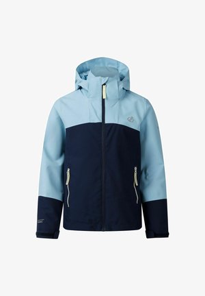 Jonge persoon draagt een blauw en marineblauw jack met marineblauwe korte broek, houdt een blauwgroene tas vast, staand tegen een effen lichte achtergrond.