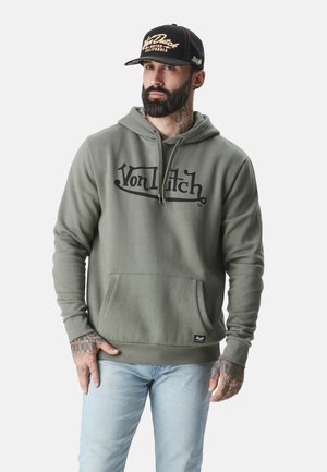 Homme avec barbe et tatouages portant un sweat à capuche gris Von Dutch et une casquette noire Von Dutch, debout les mains près des poches devant un fond uni.