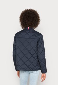 Quilted navy jakke med høj krave, der har en stribet detalje ved halsen. Materialet er glansfuldt, og designet inkluderer skrå syninger.