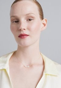 Χρυσή αλυσίδα με πέντε μικρά μαργαριτάρια, φορεμένη σε ένα ανοιχτό κίτρινο πουκάμισο με κλασικό γιακά, σε ουδέτερο φόντο.