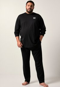 Hombre de pie descalzo, vistiendo una camiseta de manga larga negra con el texto "RELAX + CHILL" y pantalones negros a juego, frente a un fondo sencillo.