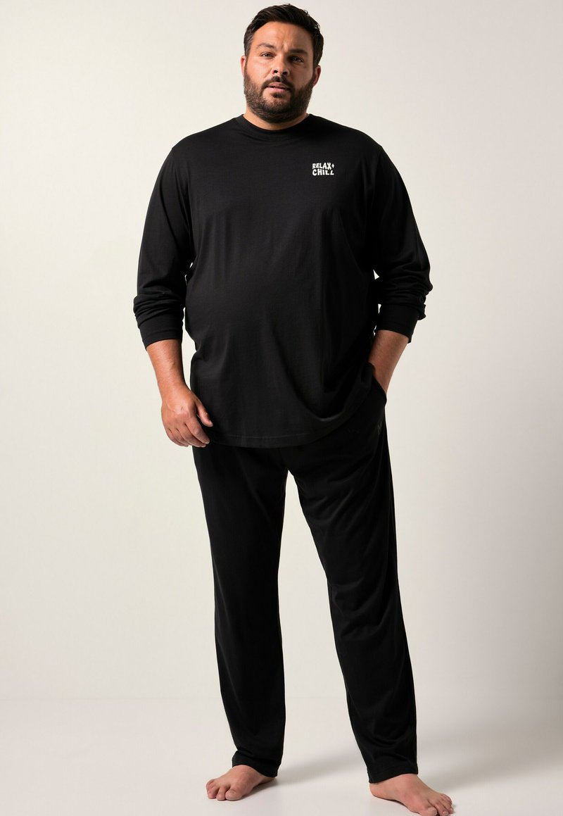 Hombre de pie descalzo, vistiendo una camiseta de manga larga negra con el texto "RELAX + CHILL" y pantalones negros a juego, frente a un fondo sencillo.