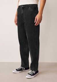 Schwarze Denim-Jeans mit einem geraden Beinschnitt, ausgestattet mit frontalen Taschen, sichtbaren Nähten und einem Knopfverschluss. Kombiniert mit schwarzen Sneakers.