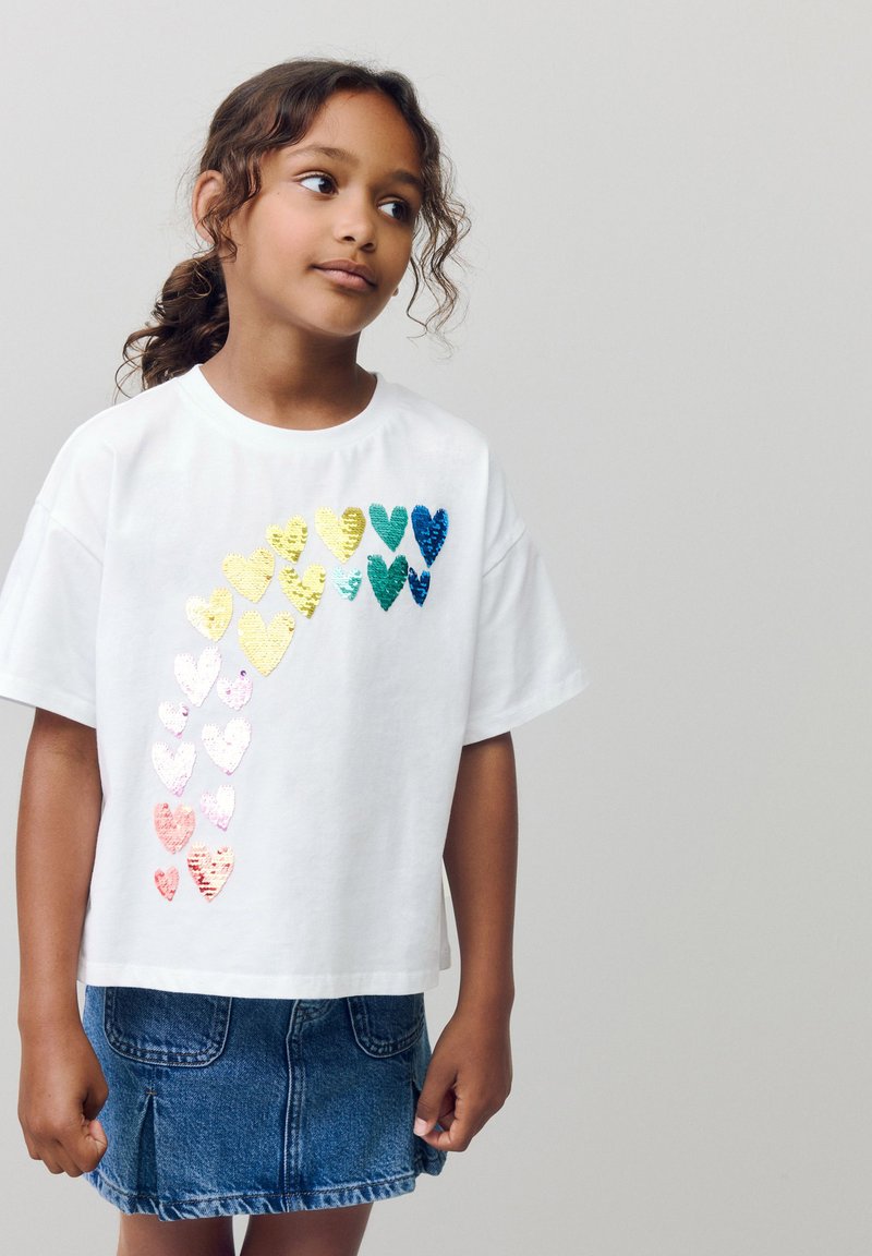 T-shirt blanc à manches courtes présentant un dégradé de cœurs en sequins colorés et brillants disposés en diagonale. Associé à une jupe en denim.