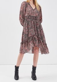 Robe à manches longues noire et rose à motifs, avec un col en V, un corsage ajusté et un ourlet en volants superposés. Portée avec des bottines noires.