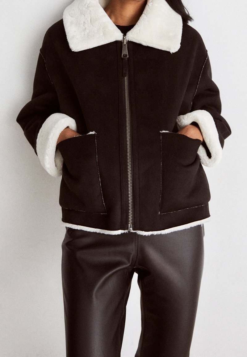 En person bär en svart jacka med vit pälskrage och manschetter, i kombination med svarta byxor, stående mot en neutral bakgrund.