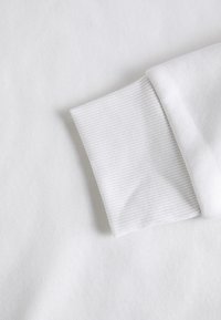 Tessuto bianco e testurizzato con polsini a coste. Il design è semplice e uniforme, con una superficie liscia e linee pulite.