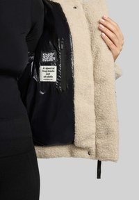 Cremefarbener Fleece-Jacke mit schwarzem Nylonfutter. Verfügt über eine mit Reißverschluss gesicherte Tasche und Etiketten, die im Futter eingenäht sind.