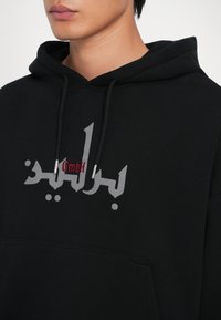 Juodas gobtuvas su raišteliu, išsiskiriantis ryškiu pilku logotipu ir raudona akcentine tekstu. Minkšta tekstūra, laisvas fasonas, kengūros tipo kišenė.