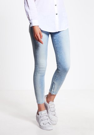 Personne portant un jean skinny bleu clair avec des fermetures éclair aux chevilles, une chemise blanche boutonnée et des baskets blanches avec trois bandes velcro.